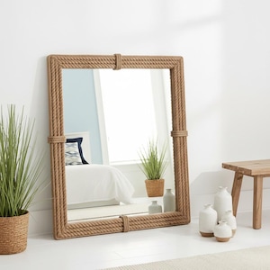 Op de afbeelding: Een grote rechthoekige spiegel met een dik touwframe. De spiegel leunt tegen een witte muur. Het frame is gemaakt van strak gewikkeld, lichtbruin touw. De spiegel weerspiegelt een slaapkamer met een bed en decoratieve items.