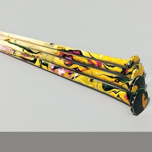 May include: A collection of decorative nails with yellow, green, and pink abstract designs. The nail heads are black with a gold accent. The nails are arranged on a light gray surface.