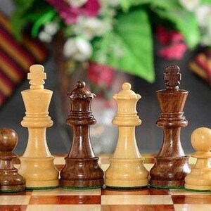 Op de afbeelding: Een schaakspel met houten stukken op een schaakbord. De stukken omvatten pionnen, een koning, een koningin en andere stukken in lichte en donkere houttinten. De achtergrond is wazig met bloemen en kussens.