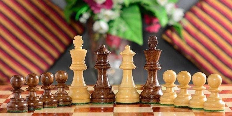 Op de afbeelding: Een schaakspel met houten stukken op een schaakbord. De stukken omvatten pionnen, een koning, een koningin en andere stukken in lichte en donkere houttinten. De achtergrond is wazig met bloemen en kussens.