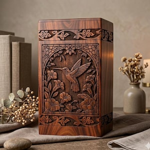 May include: A rectangular wooden box featuring detailed carvings of a hummingbird, flowers, and leaves. The box is crafted from dark brown wood. The hummingbird is depicted in flight, set within a floral and foliage design. The overall aesthetic is ornate and elegant.
