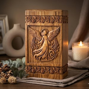 Può includere: Un'urna in legno con un'incisione di un angelo. L'urna rettangolare presenta un bordo floreale nella parte superiore e inferiore. L'angelo ha le ali spiegate ed è circondato da stelle e volute decorative. Il legno ha un tono caldo e naturale, alto circa 25 cm.