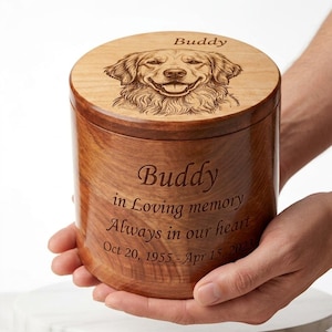 May include: A wooden pet urn with a smiling dog portrait on the lid. The urn is engraved with the name "Buddy", dates, and the inscription "in Loving memory Always in our heart". The urn is held by a person.