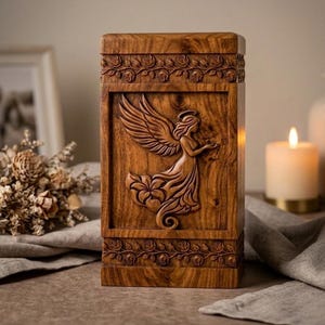 Può includere: Urna in legno con intaglio di angelo. L'urna rettangolare presenta un rilievo dettagliato di un angelo con ali, fiori e un'aureola. La parte superiore e inferiore hanno un bordo floreale. Il legno ha un tono caldo e naturale.