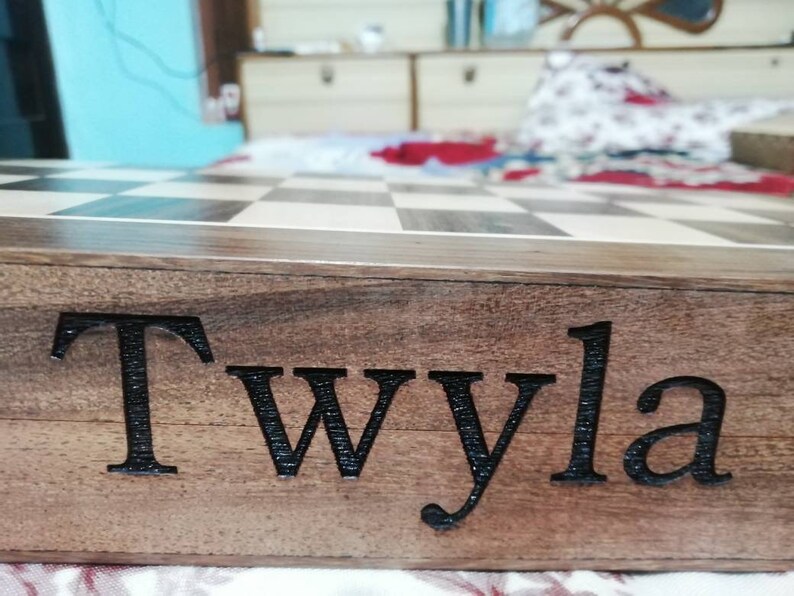 Op de afbeelding: Een houten schaakbord met een dambordpatroon. De zijkant van het bord is gegraveerd met de naam "Twyla" in zwarte letters. Het hout heeft een natuurlijke nerf.