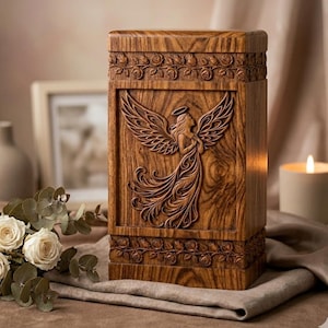 Può includere: Un'urna in legno con un disegno di angelo intagliato. L'urna rettangolare è realizzata in legno dai toni caldi e presenta un angelo con le ali spiegate sul pannello frontale. Intagli floreali adornano i bordi superiore e inferiore.