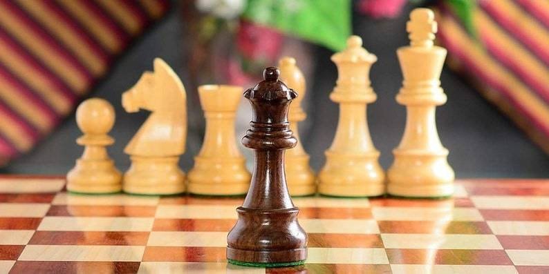 Op de afbeelding: Een close-up van een schaakspel op een schaakbord. De focus ligt op de donkerbruine koningin in de voorgrond, met andere ivoorkleurige stukken op de achtergrond. Het bord is bruin en beige.