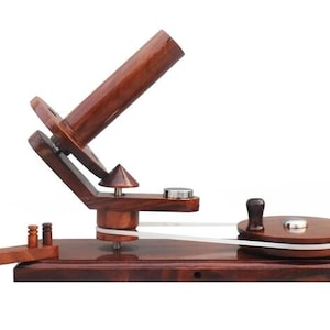 May include: A dark brown wooden yarn ball winder featuring a horizontal base, a vertical yarn guide post, a slanted spindle for holding yarn, and a circular hand-crank wheel connected by a white rubber belt to facilitate the winding process.