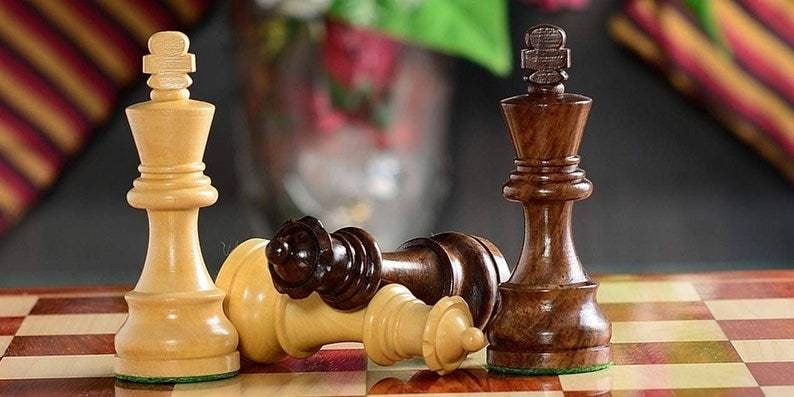 Op de afbeelding: Schaakspel met houten stukken op een schaakbord. Twee ivoorkleurige koningen staan rechtop, terwijl een donkerbruine koning en een gevallen ivoorkleurige koning op het bord liggen. De stukken zijn fijn gesneden, wat de strategie van het spel laat zien.