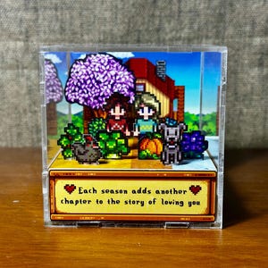 Könnte beinhalten: Ein pixeliges Diorama in einer transparenten Acrylbox. Die Szene zeigt ein Haus, Charaktere und einen Hund. Der Text unten lautet: "Jede Jahreszeit fügt ein weiteres Kapitel der Geschichte Ihrer Liebe hinzu."