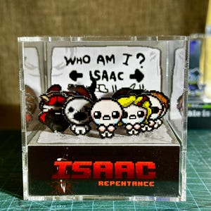 Peut inclure: Un présentoir en acrylique transparent avec des figurines pixélisées de "The Binding of Isaac: Repentance". Les figurines incluent Isaac et d'autres personnages, avec le texte "WHO AM I?" et "ISAAC" sur un fond. Le logo du jeu est en bas.