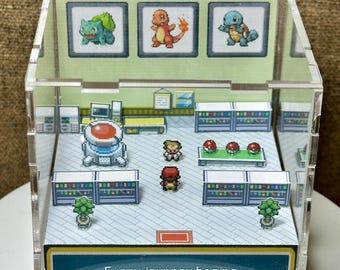 May include: A clear acrylic cube diorama featuring a miniature 2D pixel art scene of a laboratory with two characters, shelves, and a healing machine. The front panel displays the text: Every journey begins with a choice! Three framed images of Bulbasaur, Charmander, and Squirtle are mounted on the back wall.
