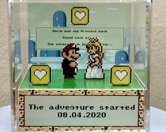 Op de afbeelding: Een heldere acrylbox met gepixelde figuren van Mario en Prinses Peach. De tekst "The adventure started 08.04.2020" is zichtbaar. De achtergrond toont gepixelde harten en een groen landschap.