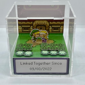 May include: A clear acrylic cube display case with a miniature 8-bit pixel art scene of Link and Zelda from the Legend of Zelda video game series. The scene depicts Link and Zelda kissing in front of a house. The base of the display case has a white card with the text "Linked Together Since 09/03/2022".