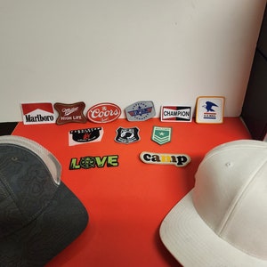 May include: Two baseball caps, one gray with a mesh back and a black brim, and one white with a black brim.  The caps are surrounded by various patches, including logos for Marlboro, Coors, High Life, Champion, and the U.S. Postal Service.