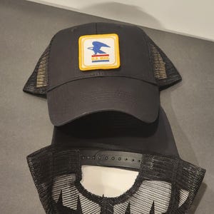 May include: Black trucker hat with a U.S. Mail patch. The patch is a square with a blue eagle, white background, and yellow border. The hat has a mesh back and a curved brim.