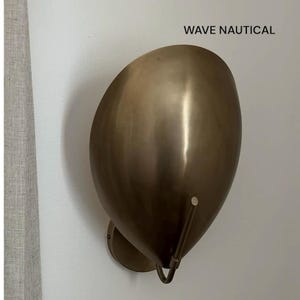 Op de afbeelding: Een messingkleurige wandlamp met een gebogen, druppelvormige kap. De lamp heeft een cirkelvormige basis en een gebogen arm die vanaf de onderkant uitsteekt. De tekst "WAVE NAUTICAL" staat erboven.