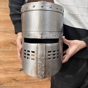 Peut inclure: Un casque de chevalier argenté avec une visière et une face avant avec un motif en grille. Le casque est doté de rivets sur les bords et d'une surface texturée.