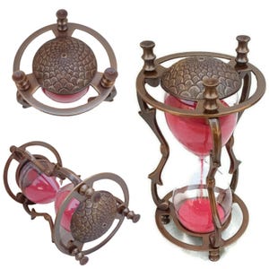 May include: A vintage-style hourglass with a bronze metal frame. The hourglass contains red sand and has a decorative, textured top and bottom. The design features a circular frame and ornate details, showcasing a classic aesthetic.
