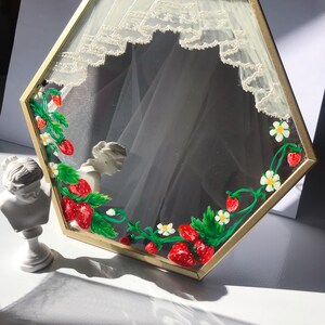 Puede incluir: Un espejo hexagonal con marco de metal dorado, decorado con fresas rojas, hojas verdes y flores blancas pintadas a mano en los bordes, junto con un dise&ntilde;o de cortina de encaje blanco pintado en la parte superior del cristal.