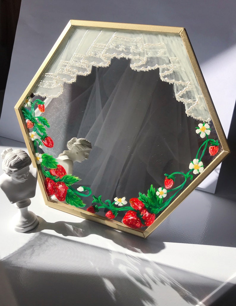Puede incluir: Un espejo hexagonal con marco de metal dorado, decorado con fresas rojas, hojas verdes y flores blancas pintadas a mano en los bordes, junto con un dise&ntilde;o de cortina de encaje blanco pintado en la parte superior del cristal.