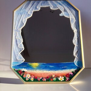 Peut inclure: Miroir octogonal avec un cadre doré. Le bord du miroir présente une scène peinte d'une plage avec de l'eau bleue, un coucher de soleil et des fleurs colorées. Au-dessus, des rideaux bleus peints avec une bordure blanche encadrent la surface réfléchissante du miroir.