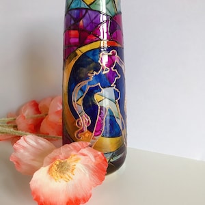 Puede incluir: Un jarrón de vidrio con un diseño de vitral que presenta una luna y una figura en un esquema de color azul y morado. El jarrón está decorado con detalles dorados.