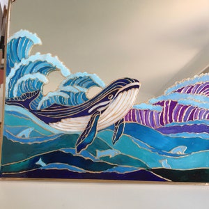 Puede incluir: Un espejo cuadrado con un dise&ntilde;o pintado a mano de una gran ballena nadando entre olas oce&aacute;nicas. La obra utiliza tonos azules, morados y blancos con contornos dorados, representando una ballena, olas rompientes y siluetas de peces.