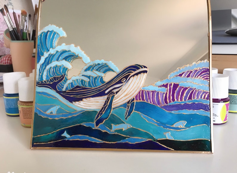Puede incluir: Un espejo cuadrado con un dise&ntilde;o pintado a mano de una gran ballena nadando entre olas oce&aacute;nicas. La obra utiliza tonos azules, morados y blancos con contornos dorados, representando una ballena, olas rompientes y siluetas de peces.