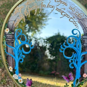 Puede incluir: Un espejo redondo con marco dorado que presenta detalles pintados a mano, incluyendo dos &aacute;rboles de color azul brillante, peque&ntilde;as flores blancas y rosas, hojas verdes y dos mariposas moradas. La parte superior muestra una escritura dorada.