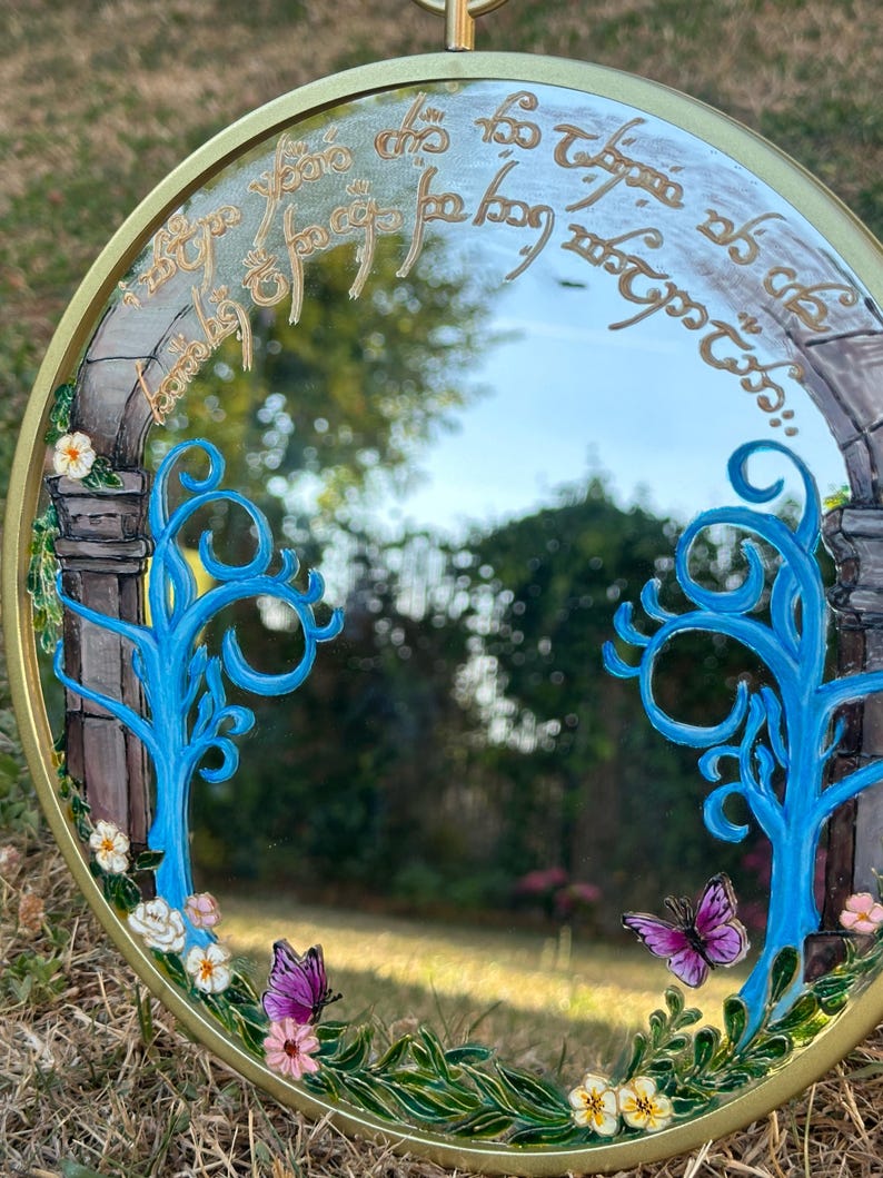 Puede incluir: Un espejo redondo con marco dorado que presenta detalles pintados a mano, incluyendo dos &aacute;rboles de color azul brillante, peque&ntilde;as flores blancas y rosas, hojas verdes y dos mariposas moradas. La parte superior muestra una escritura dorada.