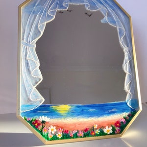 Puede incluir: Un espejo octogonal con marco dorado, pintado a mano con una escena de playa al atardecer. El dise&ntilde;o presenta cortinas azules transparentes con borde de cuentas blancas, un oc&eacute;ano azul, una playa de arena y flores silvestres.