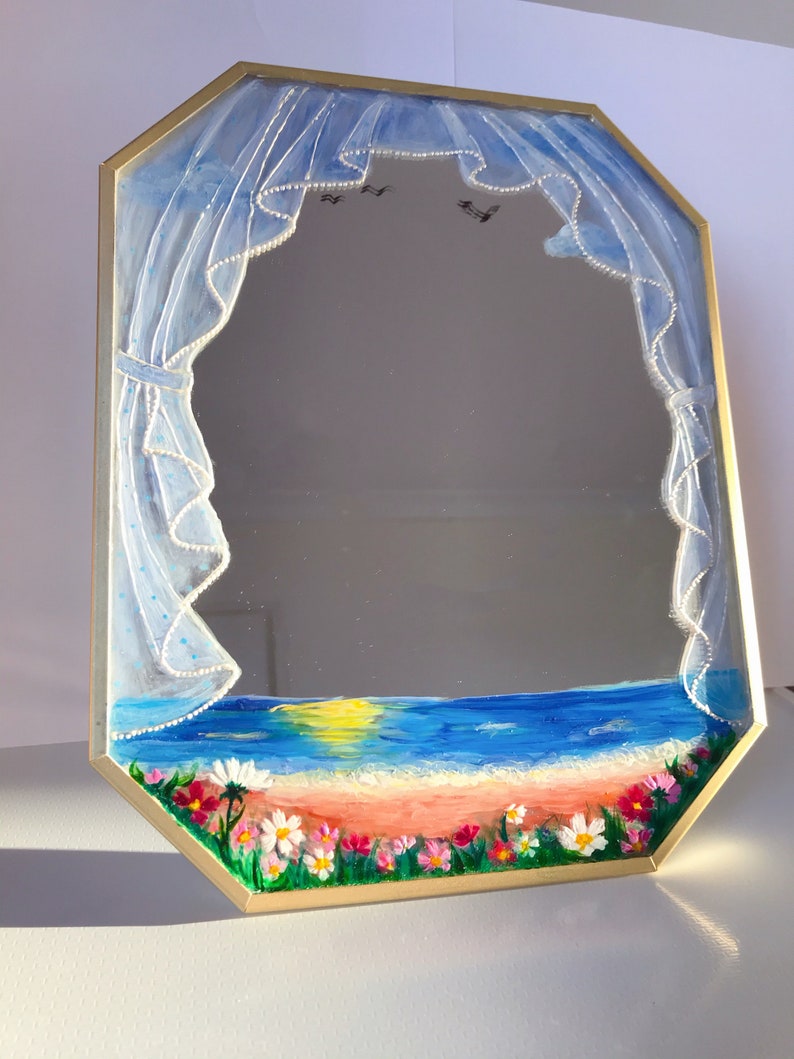 Puede incluir: Un espejo octogonal con marco dorado, pintado a mano con una escena de playa al atardecer. El dise&ntilde;o presenta cortinas azules transparentes con borde de cuentas blancas, un oc&eacute;ano azul, una playa de arena y flores silvestres.