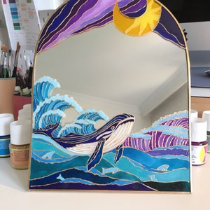 Puede incluir: Un espejo arqueado con un dise&ntilde;o pintado a mano de una ballena nadando entre olas azules y moradas. La parte superior del espejo muestra una luna creciente amarilla sobre un cielo morado oscuro. Todo el dise&ntilde;o est&aacute; delineado en color dorado.