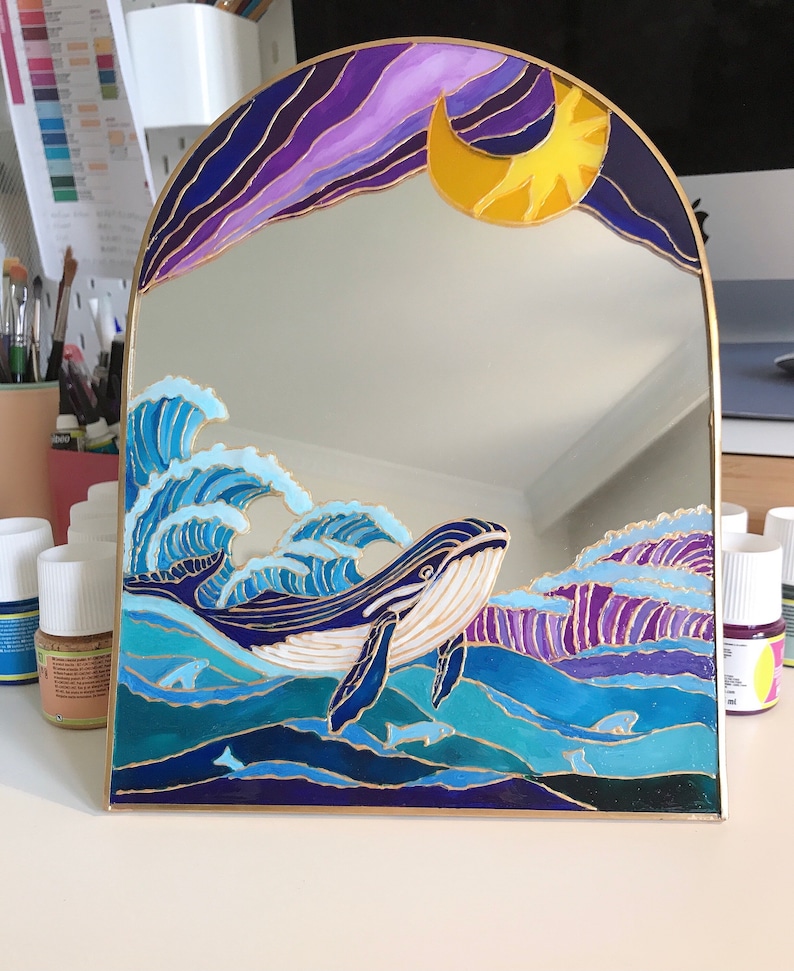 Puede incluir: Un espejo arqueado con un dise&ntilde;o pintado a mano de una ballena nadando entre olas azules y moradas. La parte superior del espejo muestra una luna creciente amarilla sobre un cielo morado oscuro. Todo el dise&ntilde;o est&aacute; delineado en color dorado.