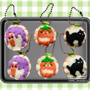 May include: A collection of Halloween-themed felt ornaments. The ornaments feature designs of a purple monster, an orange jack-o'-lantern, and a black cat. Each ornament is round, with a white base and a silver chain for hanging.