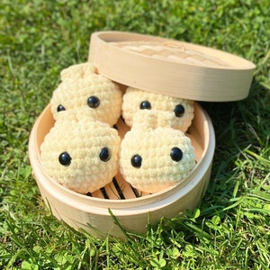 May include: A set of five crocheted stuffed buns in a bamboo steamer. The buns are cream colored with black button eyes.