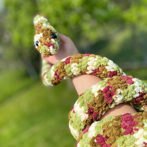 May include: A crocheted snake toy with a camouflage pattern in shades of green, brown, and burgundy. The snake is curled up and has a black eye.