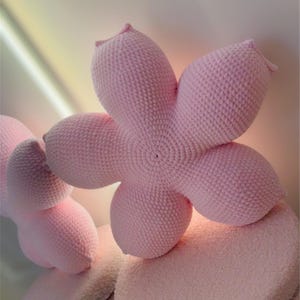 Könnte beinhalten: Nahaufnahme eines hellrosa, gehäkelten, blütenförmigen Kissens. Das Kissen hat fünf Blütenblätter und liegt auf einem passenden, runden, strukturierten Kissen. Das weiche, plüschige Design ist ideal für die Heimdekoration.