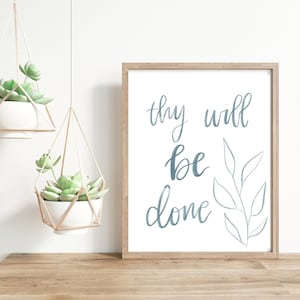 May include: A framed print with a white background and a blue handwritten quote that reads "thy will be done". A simple line drawing of a branch with leaves is to the right of the quote. The print is hanging on a white wall with two hanging plants in the foreground.