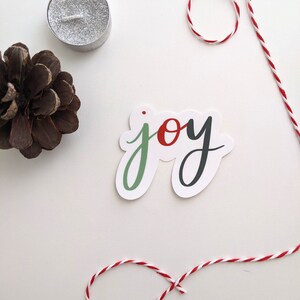 Może przedstawiać: Biała naklejka z napisem "joy" napisanym zielonym, czerwonym i czarnym pismem. Naklejka znajduje się na białym tle z czerwoną i białą sznurkiem.