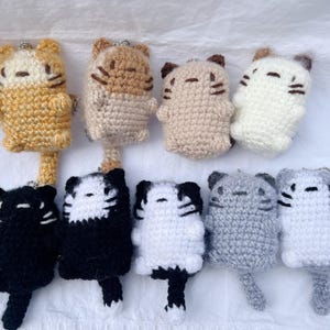 Puede incluir: Una colección de nueve llaveros de gato tejidos a ganchillo en varios colores, incluyendo naranja, beige, blanco, negro y gris. Cada figura de peluche presenta detalles faciales bordados, orejas pequeñas y una cola, con herrajes metálicos.