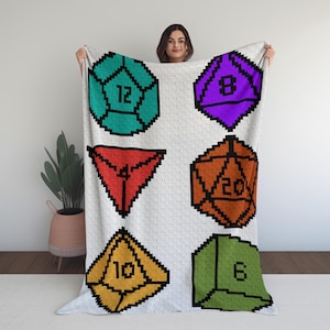 May include: A white blanket featuring pixelated dice in various colors: teal, purple, red, orange, yellow, and green. Each die displays a different number, including 4, 6, 8, 10, 12, and 20. The blanket is held up by a person.