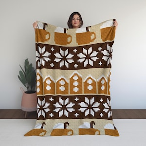May include: A warm blanket with a repeating pattern of pixelated coffee mugs, snowflakes, and gingerbread houses in brown, white, and beige. The blanket is held up by a person.