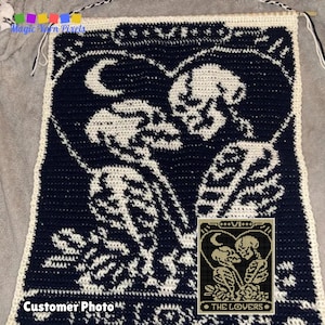 Könnte beinhalten: Ein Häkelmuster der Tarotkarte der Liebenden, mit zwei Skeletten, die sich in Herzform umarmen. Das Muster ist in schwarzer und weißer Wolle gearbeitet, wobei die Skelette weiß und der Hintergrund schwarz sind. Die Karte ist unten rechts mit "VI DIE LIEBENDEN" beschriftet.
