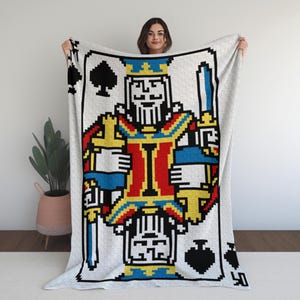 May include: A large, rectangular blanket featuring a pixelated design of the King of Spades playing card. The card's design includes a king in a crown, holding a sword, with red, blue, yellow, and black details. The blanket is held up by a person.