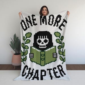 May include: A white blanket with the text "ONE MORE CHAPTER" in black pixelated font. The blanket features a pixelated illustration of a skull reading a green book, surrounded by green leaves. The blanket is held up by a person.