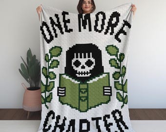 May include: A white blanket with the text "ONE MORE CHAPTER" in black pixelated font. The blanket features a pixelated illustration of a skull reading a green book, surrounded by green leaves. The blanket is held up by a person.