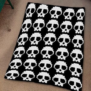 May include: A black and white crocheted blanket featuring a repeating pattern of pixelated skulls. The skulls are white against a black background. The blanket is rectangular and appears to be made of soft yarn.