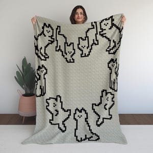 Peut inclure: Une couverture gris clair avec un motif répété de petits chats noirs pixélisés. Les chats sont disposés en cercle sur le bord. La couverture est tenue par une personne.